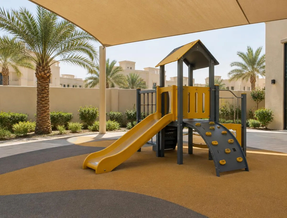 Shaded outdoor play area with a small yellow slide, a climbing frame, soft safety flooring and palm trees
